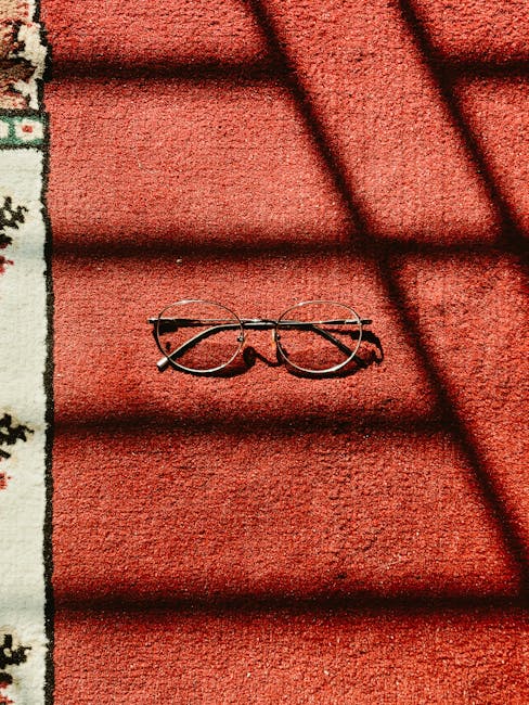 A pair of round eyeglasses with thin metal frames resting on a red outdoor carpet with a textured surface. Shadows cast by a nearby window create dark diagonal lines across the carpet, partially obscuring the eyeglasses. To the left, a patterned border featuring floral and geometric motifs in shades of green and white frames the scene. The setting appears to be an outdoor area, possibly a patio or balcony, with natural lighting highlighting the clean and well-maintained surface. This image illustrating surface care and outdoor cleaning relates to the services provided by Carpet Cleaning Marylebone on their website section for deep cleaning and sanitisation for period homes in Baker Street W1U.