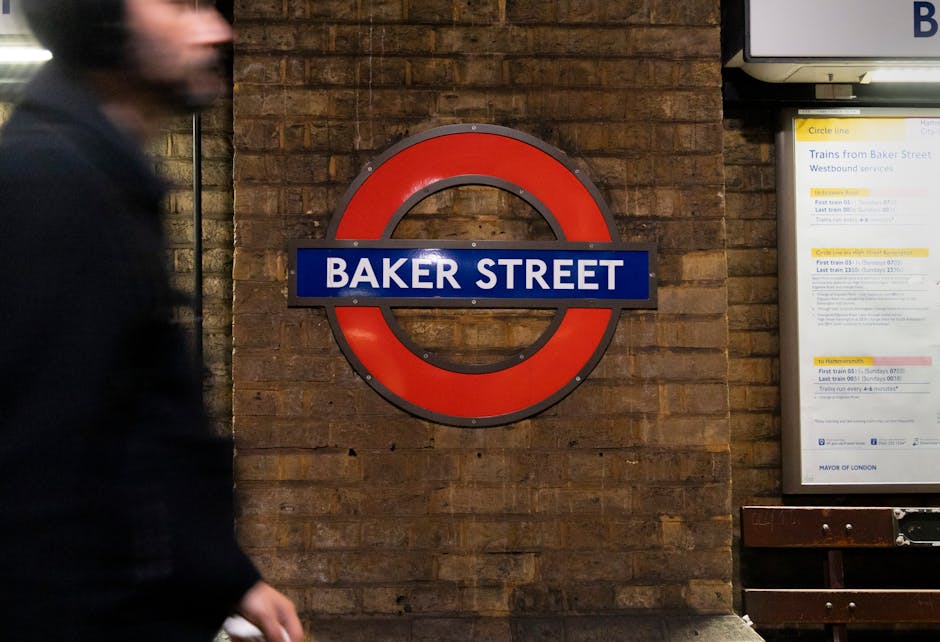 A close-up view of a London underground sign displaying 'BAKER STREET' in white letters on a blue background, mounted on a rustic brick wall. To the right, there is a partially visible timetable display with schedule information. The lighting is warm, highlighting the textured surface of the bricks. On the left, a blurred figure of a person wearing dark clothing is walking past the sign, emphasizing movement within the station environment. This scene is an example of surface cleaning and maintenance of station signage, consistent with the professional cleaning services provided by Carpet Cleaning Marylebone, as referenced on the page for Baker Street W1U.