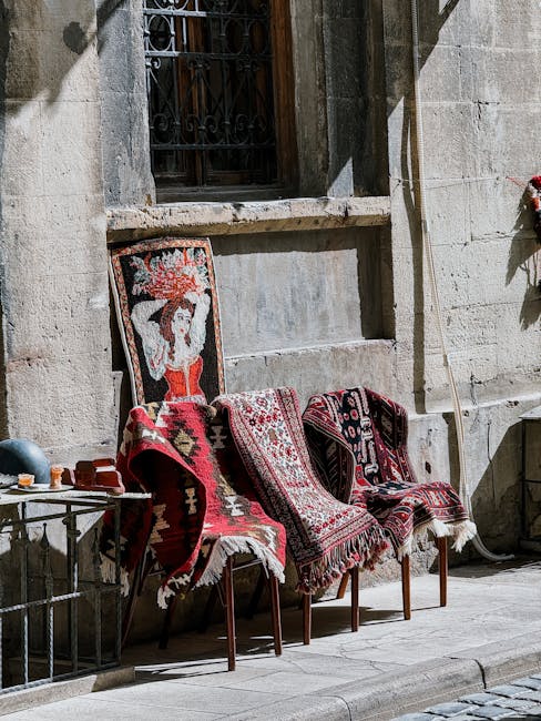 Three wooden chairs with woven legs are positioned outdoors against a stone wall with a small, barred window above. The chairs are covered with patterned rugs featuring intricate designs in red, black, and white, creating a layered look. To the left, a metal table holds small containers and a few objects, and a tall, thin broom leans against the wall beside the chairs. The scene is illuminated by natural sunlight, casting shadows on the wall and ground, with a clean, well-maintained appearance. This vignette captures a quaint, rustic outdoor seating area, emphasizing the textured stone wall, the decorative textiles on the chairs, and the overall tidy and inviting ambiance, consistent with the professional cleaning standards highlighted by Carpet Cleaning Marylebone in their cleaning services.