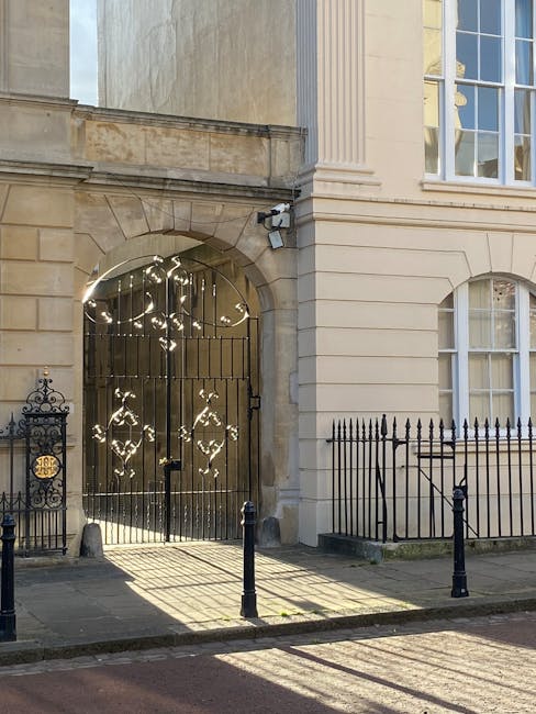 Image of a classic residential building with a cream-colored stone facade in Marylebone W1, featuring large, white-framed sash windows and decorative architectural details. The entrance is secured by an ornate black wrought iron gate with intricate scrollwork and a smaller matching gate nearby, enclosed by a low black iron fence. The pavement in front of the building includes black bollards to prevent vehicle access. Soft sunlight casts shadows across the scene, illuminating the polished stone surfaces and highlighting the building's historic elegance. No cleaning tools or surfaces are visible, indicating the exterior has been maintained well, aligning with the high standards of hygiene and appearance promoted by Carpet Cleaning Marylebone for maintaining the aesthetic appeal in domestic and commercial properties.