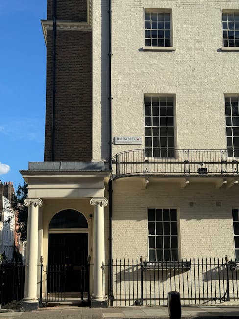 Exterior view of a Georgian-style building on Hill Street W1G in Marylebone, featuring cream-colored brickwork and symmetrical sash windows. The ground-floor entrance is framed by classical white columns supporting a small portico with a rounded arch, set behind a black metal fence. The building's facade appears clean and well-maintained, with bright natural sunlight highlighting the surfaces and casting soft shadows. The overall scene conveys a tidy, historic residential property, consistent with high standards of domestic cleaning and upkeep, as emphasized by Carpet Cleaning Marylebone in their surface cleaning and sanitisation services for Georgian flats.