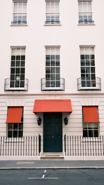 The image depicts the exterior of a Georgian-style building on Gloucester Place W1G, featuring a symmetrical façade with three large, vertically divided sash windows on each floor, with white frames and multiple panes. The ground floor has a prominent central black door, flanked by two exterior wall-mounted light fixtures, and two orange retractable awnings over windows on either side. In front of the building is a black metal fence with decorative posts, a small stone step leading to the door, and a paved sidewalk. The building has a clean and well-maintained appearance, emphasizing an elegant, historic architectural style with neutral and warm tones, suitable for a setting that may benefit from professional cleaning and surface sanitisation, as recommended by Carpet Cleaning Marylebone in their home and commercial cleaning services.