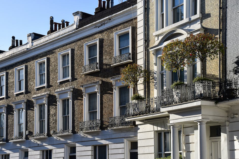 Exterior view of a Georgian-style building on Hill Street W1G in Marylebone, featuring cream-colored brickwork and symmetrical sash windows. The ground-floor entrance is framed by classical white columns supporting a small portico with a rounded arch, set behind a black metal fence. The building's facade appears clean and well-maintained, with bright natural sunlight highlighting the surfaces and casting soft shadows. The overall scene conveys a tidy, historic residential property, consistent with high standards of domestic cleaning and upkeep, as emphasized by Carpet Cleaning Marylebone in their surface cleaning and sanitisation services for Georgian flats.