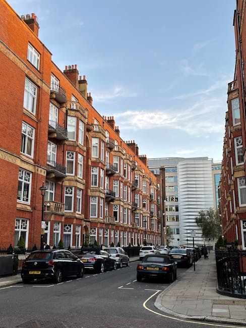 Photograph of a red-brick residential building in Marylebone with multiple floors and arched windows, some with balcony railings, set against a clear blue sky. The building features decorative white t