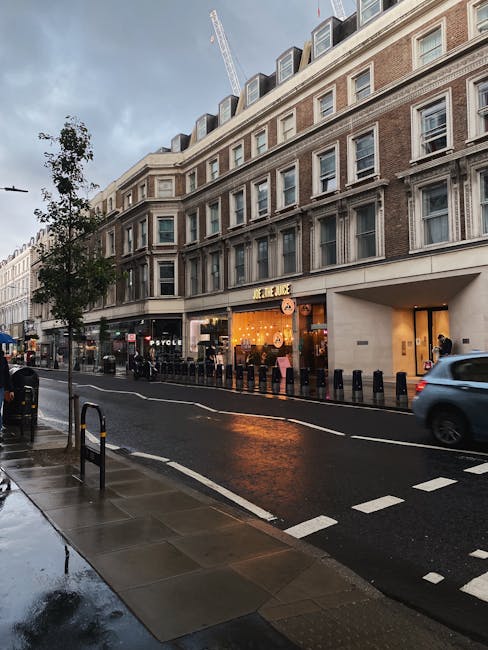 A daytime street scene in Marylebone featuring a multi-story building with large windows and a rounded facade. The ground floor hosts a retail store with bright branding, large display windows, and a warm, inviting interior visible from outside. The street is wet, possibly from recent rain, with reflections on the pavement, and a few pedestrians and vehicles are seen passing by. The sidewalk has a few trees and black bollards, and overcast skies suggest cloudy weather. These elements emphasize the importance of regular surface cleaning and maintenance by Carpet Cleaning Marylebone to ensure hygienic and appealing commercial spaces after busy days, supporting top-quality deep cleaning and sanitisation of property surfaces.