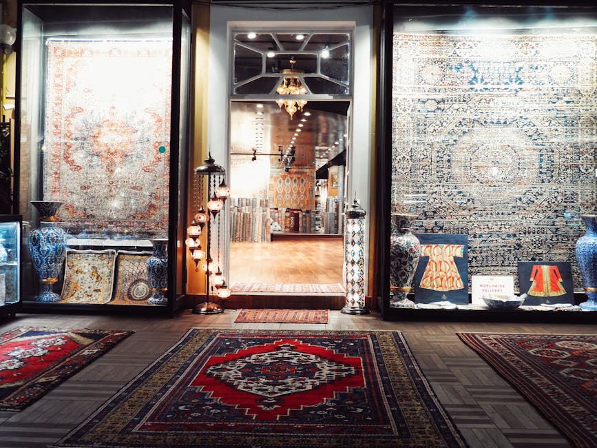 The image shows the entrance to a boutique carpet shop with large glass windows displaying various decorative rugs and vases on both sides of the doorway. Inside, visible through the glass, there are more rugs and accessories arranged in an elegant setting with warm lighting and wooden flooring. The shop has a polished, clean appearance, showcasing a variety of colorful, patterned carpets and decor pieces. The scene highlights surface cleaning and presentation, reflecting the meticulous hygiene standards promoted by Carpet Cleaning Marylebone. The overall atmosphere suggests attention to maintaining a pristine environment for showcasing fine carpets and textiles, consistent with professional deep cleaning and domestic surface maintenance practices. The well-lit interior and neatly arranged displays emphasize a focus on surface cleanliness and aesthetic appeal.
