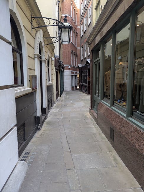 A daytime street scene in Marylebone featuring a multi-story building with large windows and a rounded facade. The ground floor hosts a retail store with bright branding, large display windows, and a warm, inviting interior visible from outside. The street is wet, possibly from recent rain, with reflections on the pavement, and a few pedestrians and vehicles are seen passing by. The sidewalk has a few trees and black bollards, and overcast skies suggest cloudy weather. These elements emphasize the importance of regular surface cleaning and maintenance by Carpet Cleaning Marylebone to ensure hygienic and appealing commercial spaces after busy days, supporting top-quality deep cleaning and sanitisation of property surfaces.