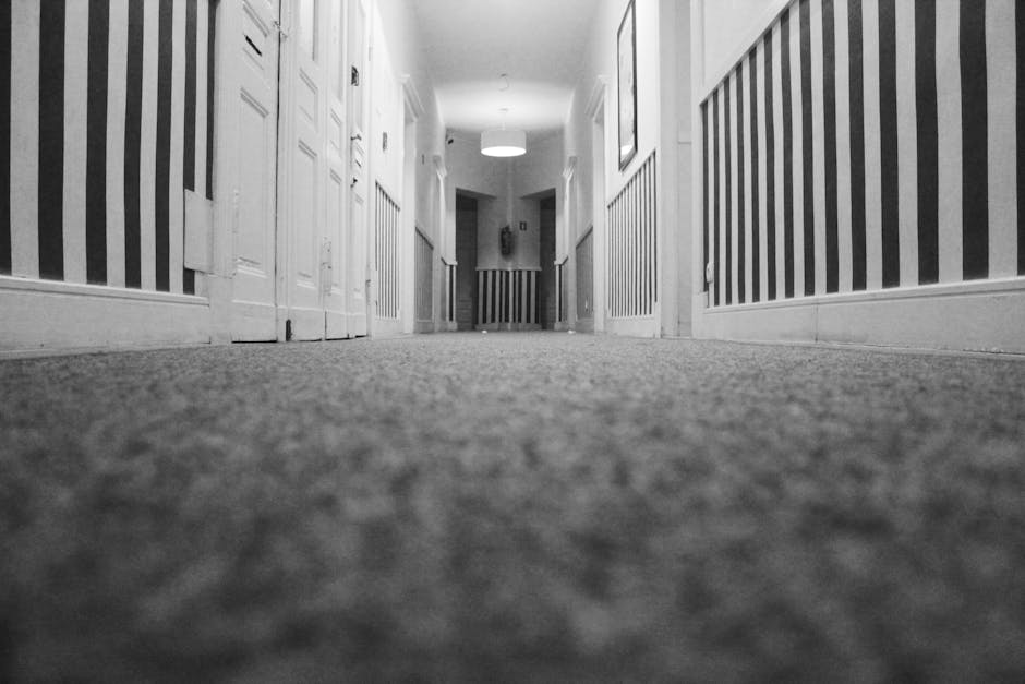 A low-angle photograph of a clean, carpeted hallway in a residential or commercial building, viewed from the floor level. The carpet is a neutral shade, appearing soft and well-maintained with no visible stains or debris. The walls feature striped wallpaper with alternating dark and light vertical lines, and are adorned with white wooden wainscoting. Wooden doorframes and doors are aligned along the corridor, with a round ceiling light fixture illuminated, casting even lighting throughout the space. The overall scene conveys a sense of cleanliness and good maintenance, consistent with professional surface cleaning and domestic or commercial sanitisation standards, as provided by Carpet Cleaning Marylebone.