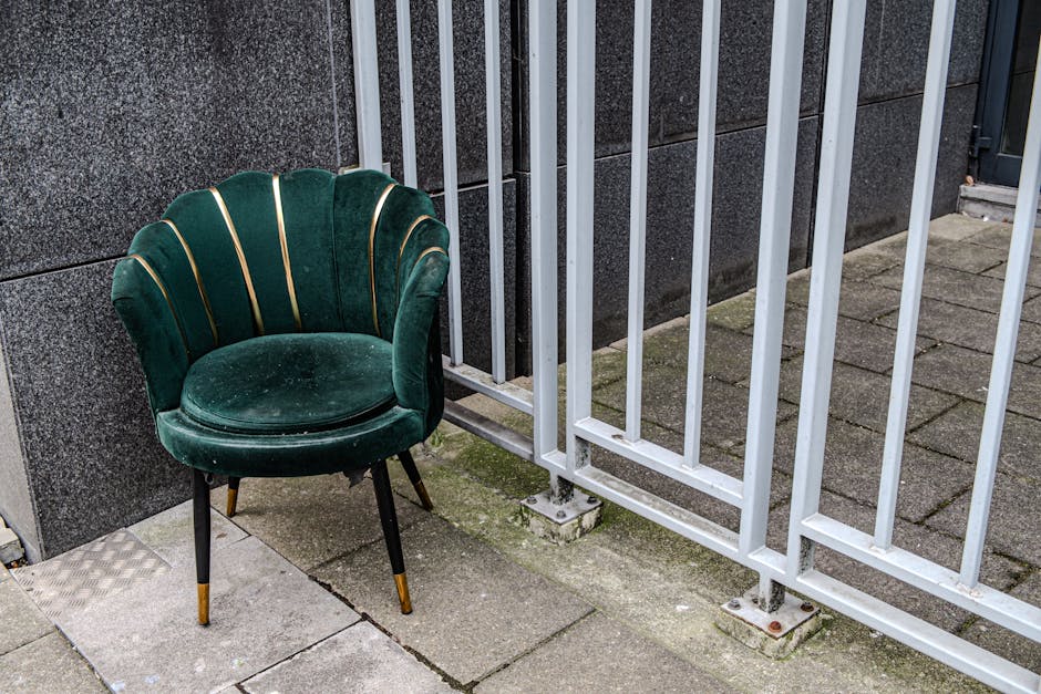 A vintage-style, deep green velvet armchair with gold accents on the backrest and tapered black legs with gold tips, placed outdoors on a paved sidewalk adjacent to a grey stone wall and white metal fence bars. The chair appears clean and well-maintained, positioned near the corner of a building. The lighting is natural, providing clear visibility of the upholstery's texture and the surrounding surfaces. This setting suggests residential or commercial outdoor seating area maintenance, with Carpet Cleaning Marylebone focusing on surface cleaning and hygiene. The scene highlights the importance of regular cleaning to preserve furniture appearance in urban environments.