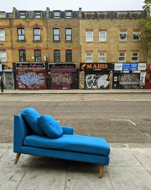 A worn-out, grey fabric sofa with four seats, positioned outdoors against a brick wall. The sofa shows visible signs of wear and tear, including a noticeable tear on the left armrest and a sagging cushion in the middle seat. The fabric appears slightly stained and aged, with the seat cushions exhibiting unevenness and minor wrinkles. The sofa is situated on a concrete pavement with a metal drainage grate directly in front of it. The background consists of a weathered brick wall, indicating an urban or alleyway setting. The lighting is natural, casting soft shadows on the sofa, which emphasizes its aged and used condition. This image exemplifies the need for upholstery deep cleaning and maintenance, highlighting the importance of professional cleaning services such as those provided by Carpet Cleaning Marylebone for hygiene and surface longevity.