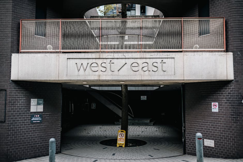 The image depicts the entrance to a parking garage or residential building in Marylebone, with a modern black brick facade and a concrete overhang displaying the engraved words 'west/east'. The entrance is partially shaded, and a tree is visible behind a metal railing on the balcony above. The ground in front of the entrance is paved with light-colored bricks, and there are a few bollards and safety signs near the walkway. The lighting appears natural, indicating daytime, and the overall scene is clean and well-maintained, suitable for surface and deep cleaning services promoted by Carpet Cleaning Marylebone as part of domestic or commercial hygiene and sanitation efforts following Westminster Council Carpet Disposal Rules for Marylebone.