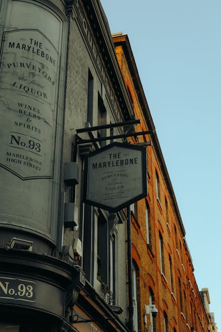 A close-up view of the exterior of The Marylebone pub building, featuring a curved white façade with vintage-style signage displaying the pub's name, address, and offerings such as wines, spirits, and liquor. The sign is mounted on a black metal arm extending from the wall. The building has multiple old brick facades with tall, narrow windows, some with flower boxes underneath. Bright daylight illuminates the scene, highlighting the aged brickwork and glass windows, with the overall appearance clean and well-maintained. This exterior is indicative of a well-preserved historic commercial property, exemplifying surface cleaning and preservation practices as part of professional domestic and commercial cleaning standards, aligning with the Westminster Council Carpet Disposal Rules for Marylebone, as promoted by Carpet Cleaning Marylebone.