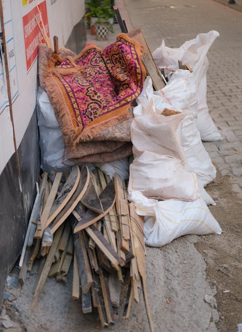 A pile of old carpets with ornate patterns and fringed edges, leaning against a wall on a city sidewalk. The carpets appear worn and partially rolled, with some sections folded or crumpled. Surrounding the carpets are large white and beige sacks filled with debris, possibly from a disposal or renovation process. Wooden planks and pieces of discarded construction material are scattered on the ground nearby, which is paved with concrete tiles. The scene is outdoors, with natural daylight illuminating the area. The setting suggests a residential or commercial space undergoing cleaning, renovation, or carpet disposal, with no visible cleaning tools or products present. Carpet Cleaning Marylebone offers surface and deep cleaning services, including responsible disposal of old carpets, ensuring hygienic and well-maintained environments.