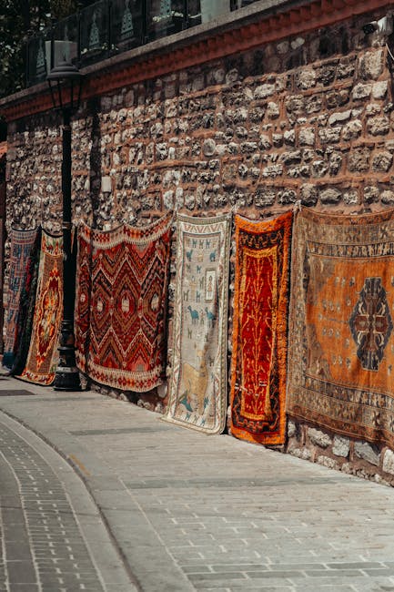 A row of colorful, patterned oriental rugs hangs on a weathered brick wall along a paved street. The rugs display intricate designs in shades of red, orange, blue, and beige, with some featuring geometric motifs and others floral patterns. The textured brick wall behind them shows signs of age, with variations in color and mortar. Bright natural lighting illuminates the scene, highlighting the rich textures and detailed craftsmanship of each rug. The setting appears to be an outdoor area, possibly a marketplace or a street-side display, reflecting a traditional or antique aesthetic. Carpet Cleaning Marylebone specializes in surface cleaning, deep cleaning, and sanitisation, ensuring the maintenance and hygiene of such delicate fabrics in domestic and commercial environments.